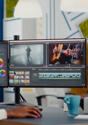 Black videographer putting on headset editing movie using post production software working in creative start up agency office. African editor processing video footage on computer with dual displays